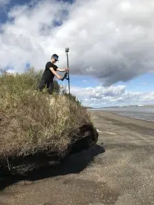 Seth Classen stands on top of a small bluff holding a Trimble instrument. The tide on the beach below is low. The sky is cloudy. 