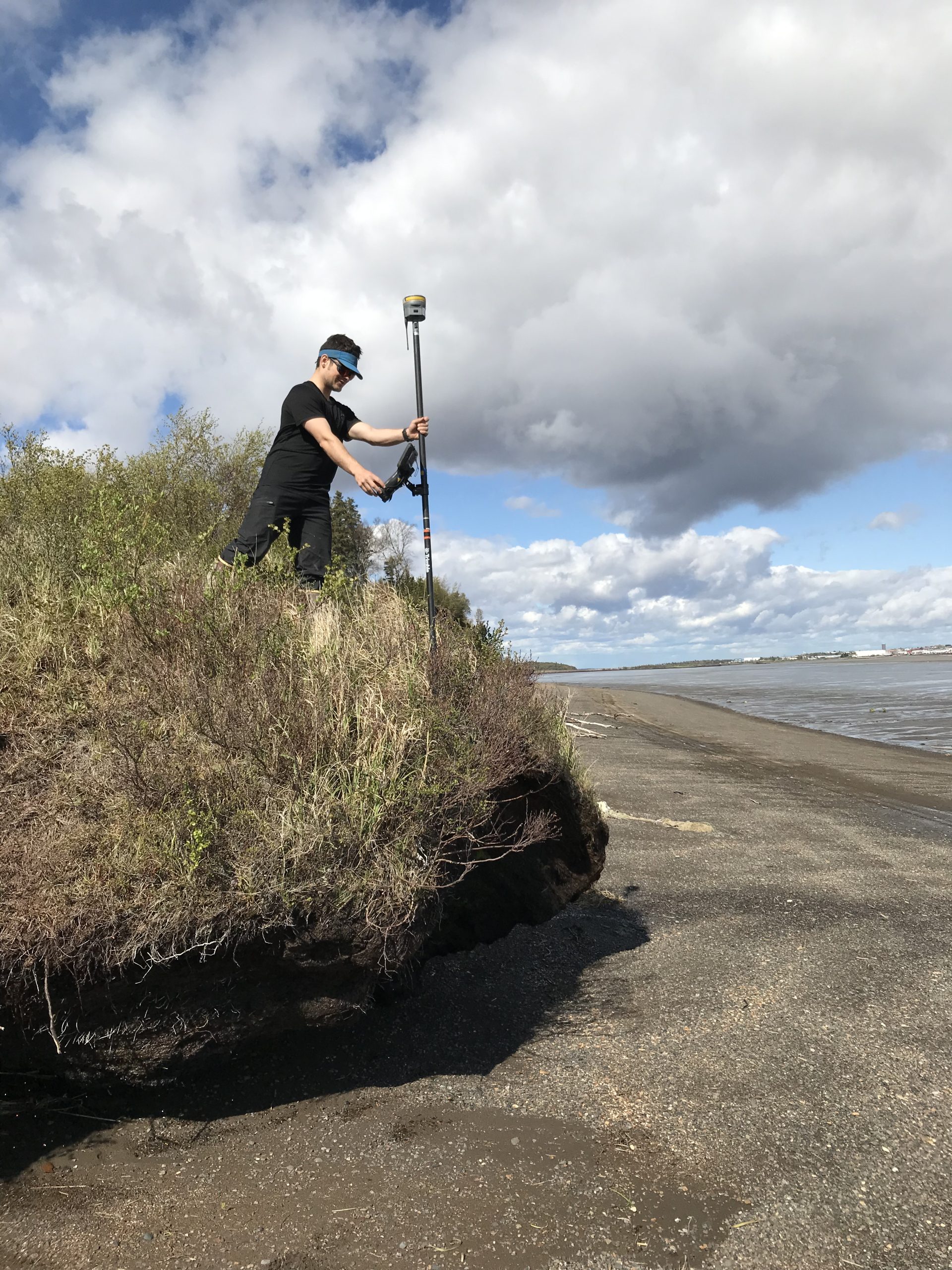 Seth Classen stands on top of a small bluff holding a Trimble instrument. The tide on the beach below is low. The sky is cloudy. 