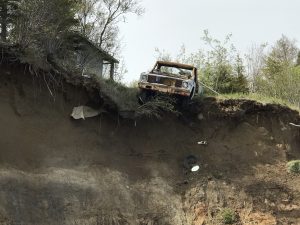 An old rusted trucks hangs halfway off the top of a tall bluff. There is grass and trees on top of the bluff. The sky is overcast.