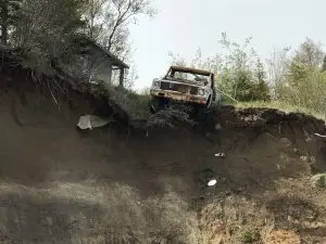An old rusted trucks hangs halfway off the top of a tall bluff. There is grass and trees on top of the bluff. The sky is overcast.