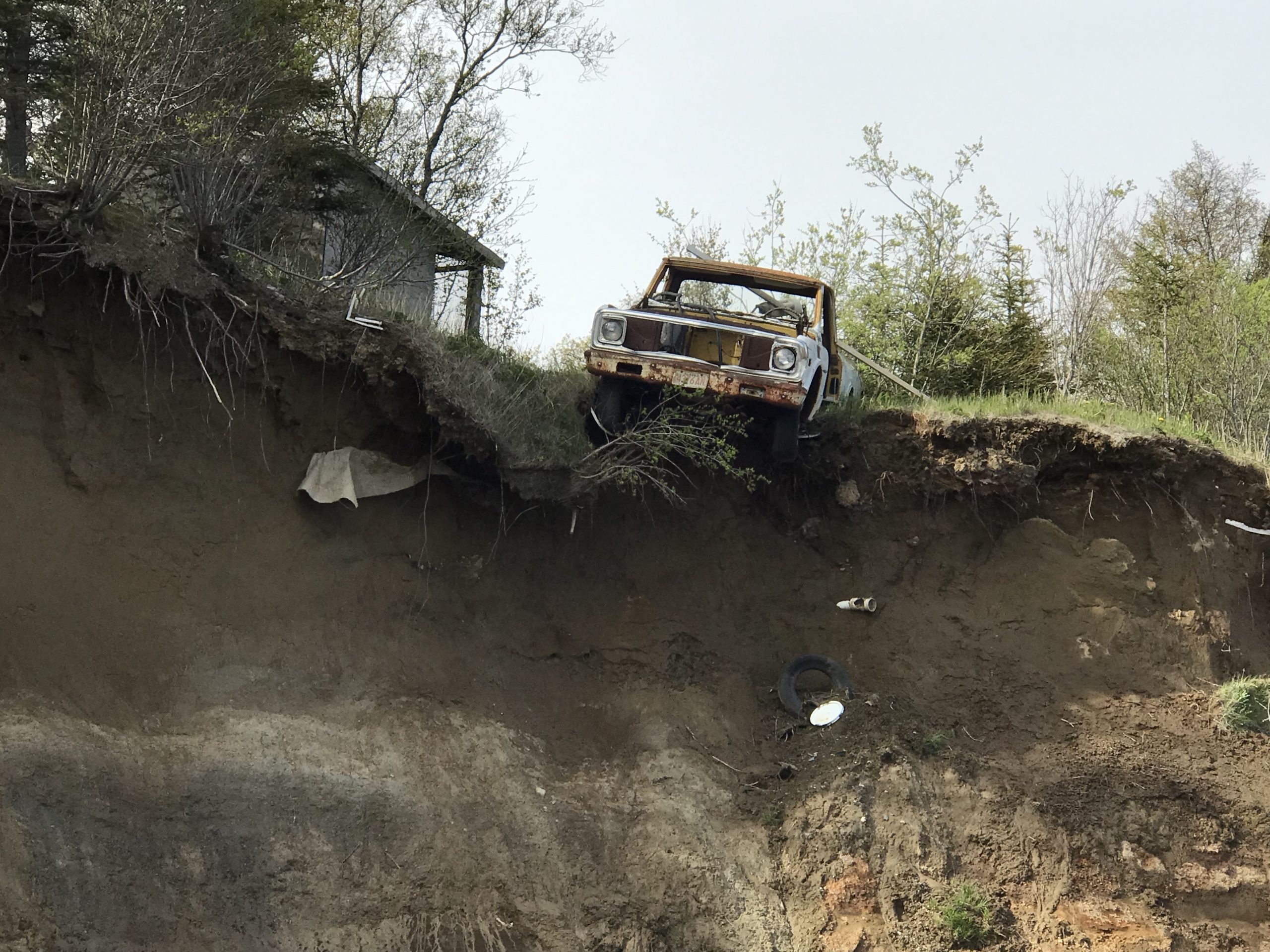 An old rusted trucks hangs halfway off the top of a tall bluff. There is grass and trees on top of the bluff. The sky is overcast.