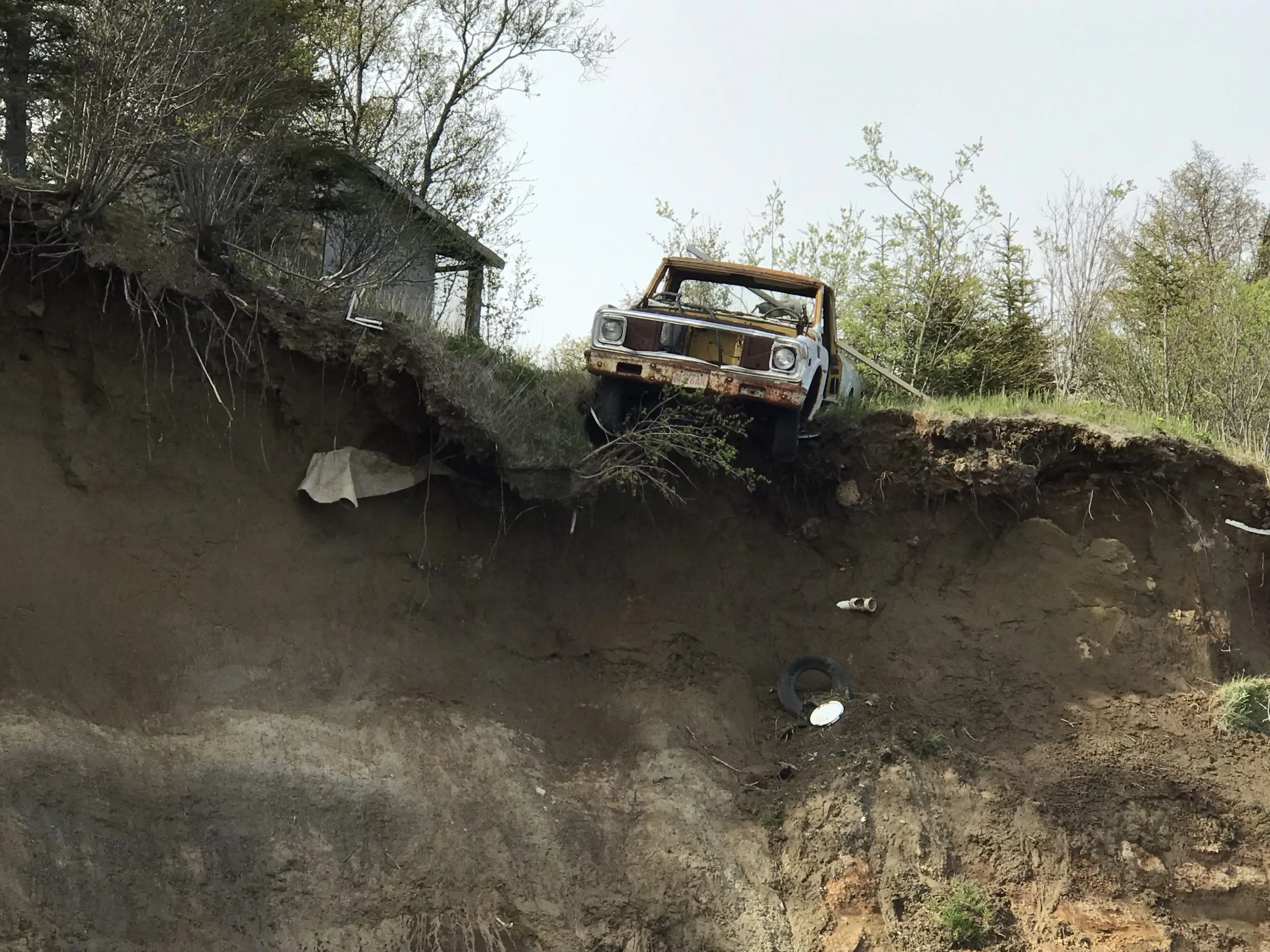 An old rusted trucks hangs halfway off the top of a tall bluff. There is grass and trees on top of the bluff. The sky is overcast.