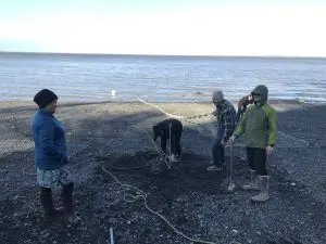 Five people stand around a rocky beach. A fishing net is being spread out and extending into the water. Someone is holding a sledge hammer. Someone else is tying down a rope. The water appears calm and the sky is bright and overcast. 