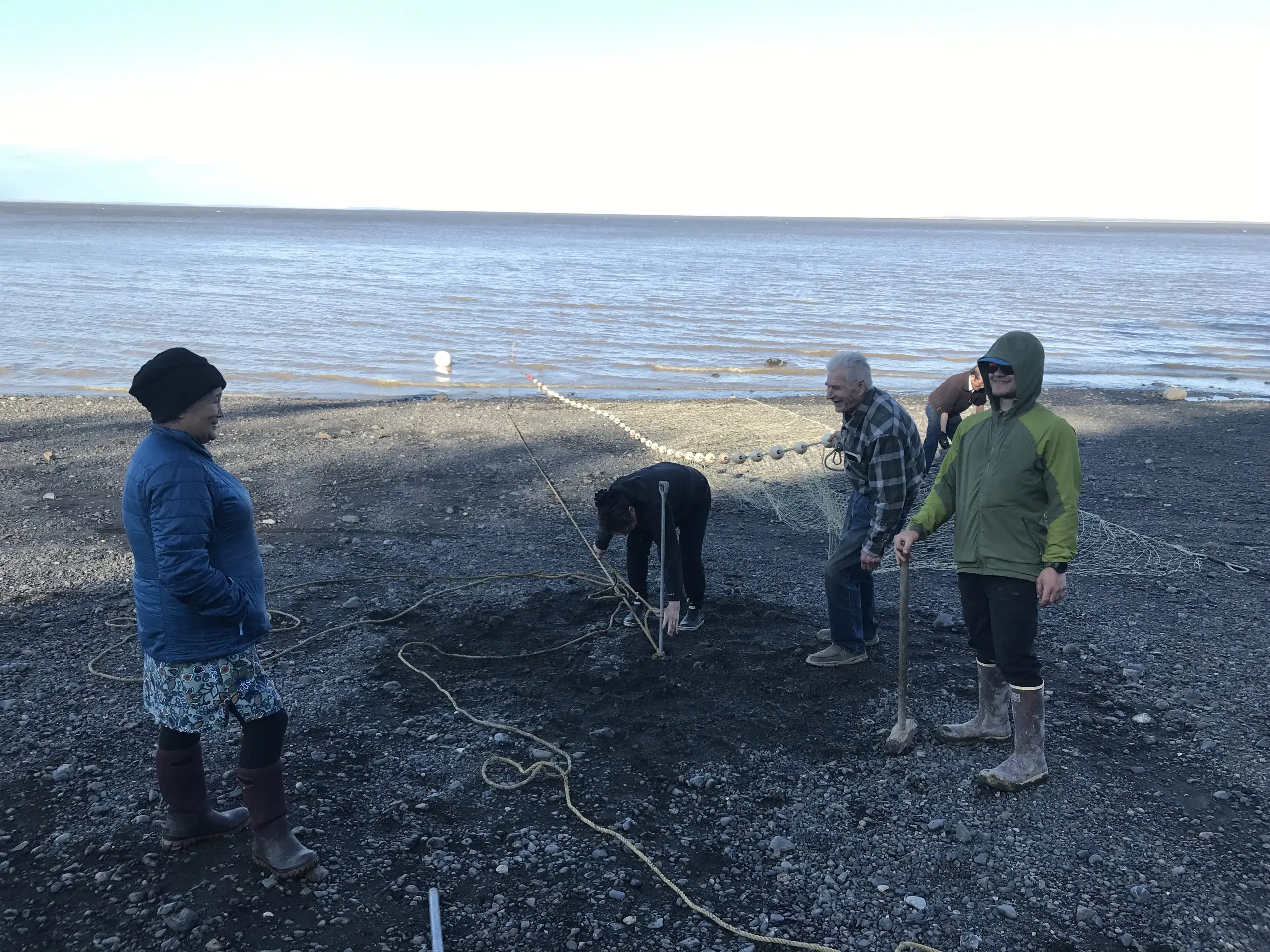 Five people stand around a rocky beach. A fishing net is being spread out and extending into the water. Someone is holding a sledge hammer. Someone else is tying down a rope. The water appears calm and the sky is bright and overcast. 