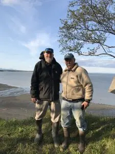 Two people stand for a photo on top of a grassy bluff. Behind them is the beach with calm ocean waters. The sky is partially cloudy. 