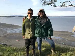 Two people standing next to each other for a photo. They are standing on top of a grassy bluff. Behind them is a low-tide beach. The sky is partially cloudy.