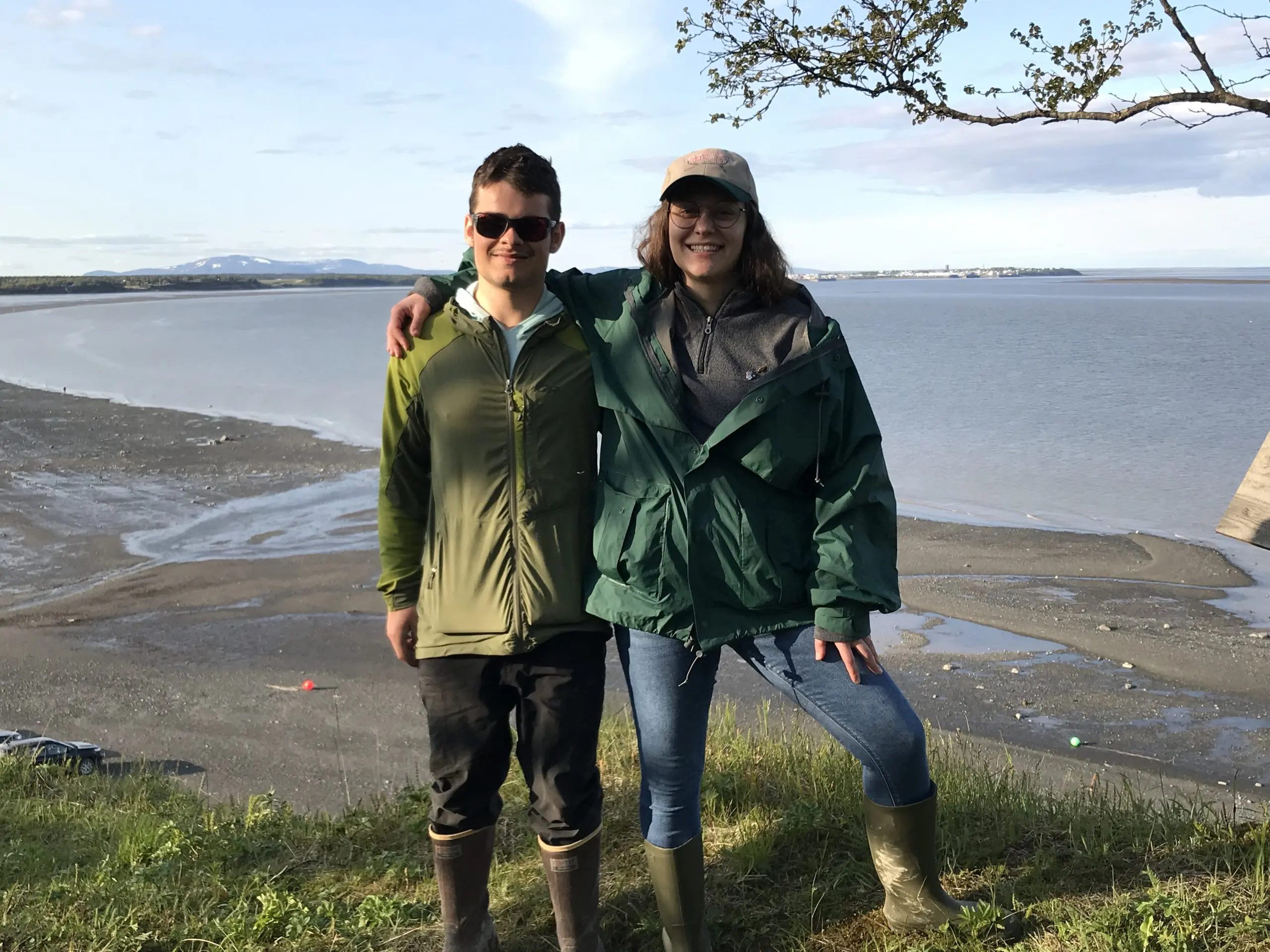 Two people standing next to each other for a photo. They are standing on top of a grassy bluff. Behind them is a low-tide beach. The sky is partially cloudy.