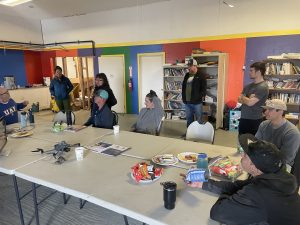 A group of people standing and sitting around tables with various snacks, papers, and a drone sat on it. The room has rainbow colors on the walls. There are some bookcases around the room. 