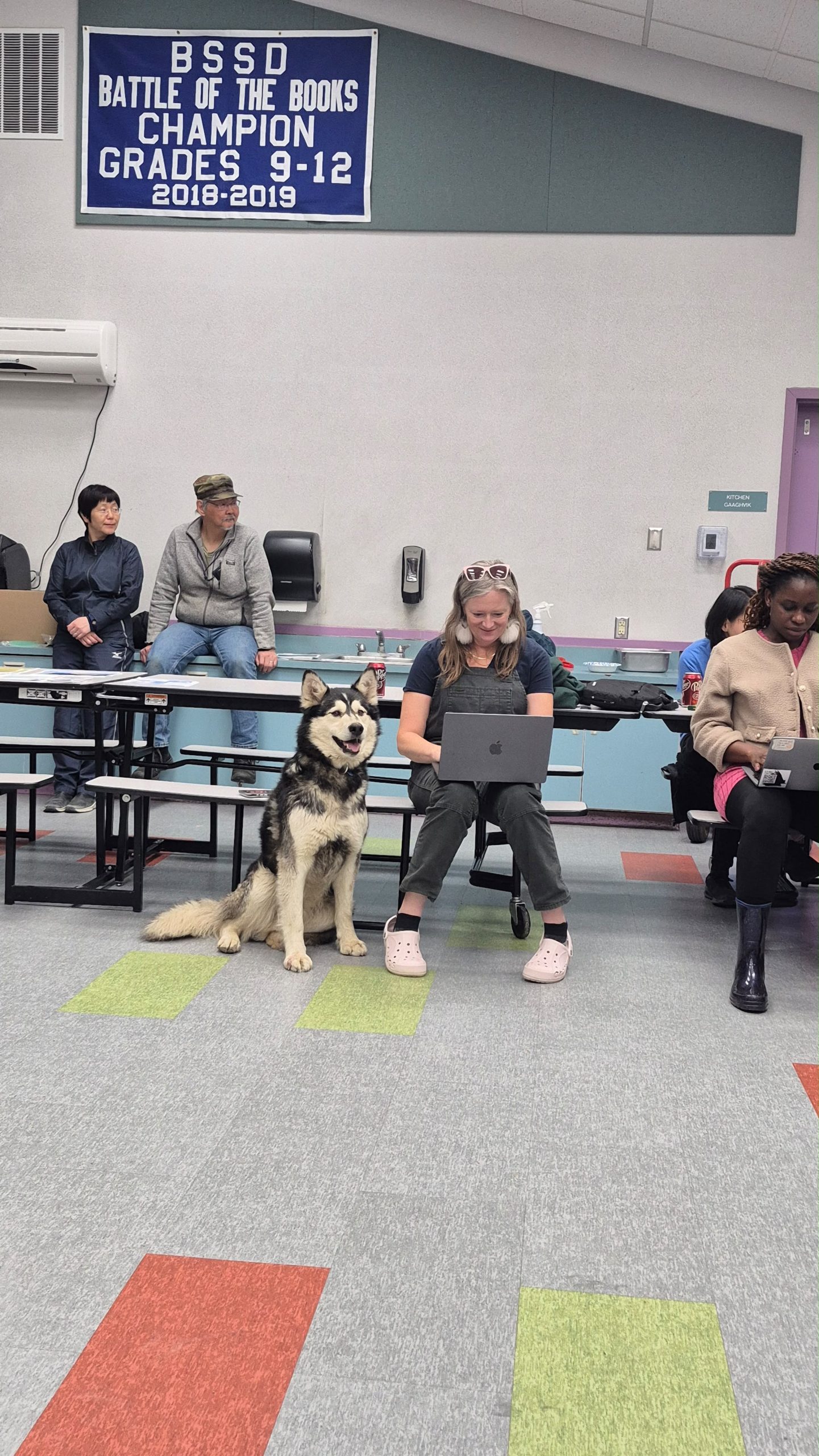 A husky is sitting next to someone sitting on a lunch table and using a laptop. There is someone next to them sitting at a different lunch table using their laptop. They appear to be in a school cafeteria. 