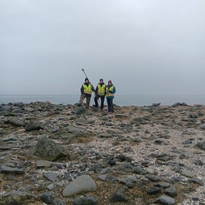 Chris Maio, Chris Tran, and Harper Baldwin stand next to each other for a photo. Chris is holding the Trimble equipment. They are standing in a rocky field. Behind them is foggy ocean waters. The sky is overcast. 