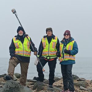 Chris Maio, Chris Tran, and Harper Baldwin stand next to each other posing for a photo. Chris Maio is holding the Trimble equipment, Chris Tran is holding the field notebook, and Harper Baldwin is holding a water bottle. They are standing in a field of rocks. Behind them is the ocean and a foggy, overcast sky.