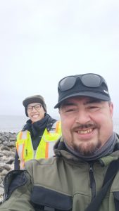 Chris and Alex stand next to each other for a selfie. They are standing in a rocky field and the sky is overcast. 
