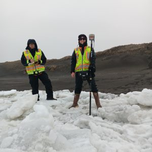 Chris and Harper stand on broken ice deposits. Harper holds the Trimble equipment while Chris is writing in the field notebook. They are on a beach. There are small hills behind them. The sky is overcast. 