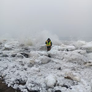 Chris Maio stands in the coastal ice deposit as a wave crashes behind him. The sky is overcast. 