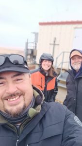 Alex, Harper, and Chris stand near each other for a selfie. There is a building behind them. The sky appears bright. 