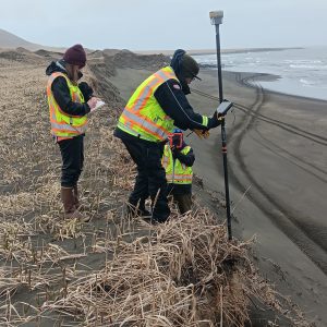Harper is taking notes in the field notebook, Chris Tran is holding the Trimble equipment, and Chris Maio is taking a photo with the field notebook. They are standing around the top of a bluff's edge with dead grass. The sky is overcast. 