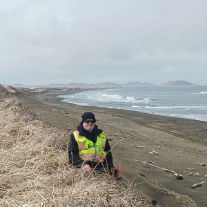 Chris sitting along the crest of a tall, sloping bluff. The top of the bluff is covered in dead grass. Chris is holding the field ipad, his arm rests on the top of the bluff. The background is a beach with some driftwood and crashing waves. The sky is overcast.