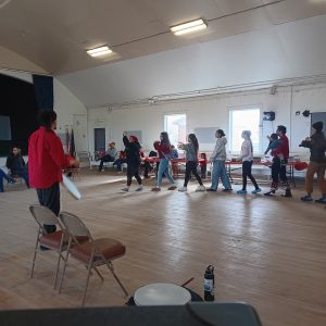A line of people walk in sync while motioning their arms, someone in the foreground is beating on a drum. They are in a gymnasium. 
