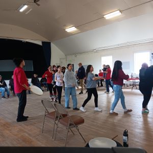 A line of people walk in sync while motioning their arms, Chris Maio is in the line joining along with the motions. They are inside a gymnasium. Someone stands off to the side holding a drum, further back people sit in folding chairs watching the dancers.