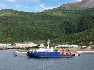 The Sikuliaq sits next to a dock on calm waters. Behind it are some buildings and foothills covered in green foliage that grows more spars the higher the hills reach. The sky can be seen in a corner of the photo with a few wispy clouds.