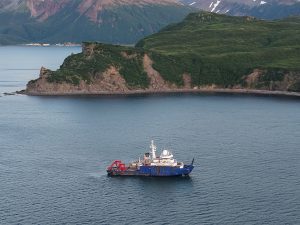 The Sikuliaq sits in the middle of the water, surrounded by cliffs that are covered in vegetation.
