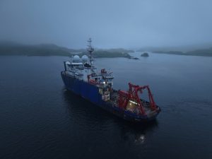 The Sikuliaq sits in the middle of calm, dark waters. There are some islands in front of it. The sky is foggy. The image seems to have been taken in the evening.