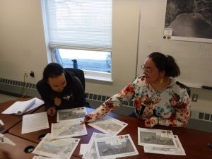 Two people sit next to each other at a table with maps spread all over. They are inside a building. There is a window behind one of them on the left of the image and a whiteboard behind the person on the right.