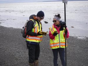 Reyce and Roberta standing next to each other on a rocky beach. Reyce is holding a field notebook and Roberta is holding the Trimble equipment. They are on a low tide beach.