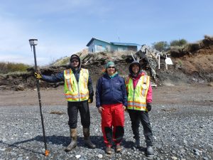Three people standing next to each other. One of them is holding the Trimble equipment. They are standing on a rocky beach. Behind them is a bluff covered in debris. There is a blue house further back. The sky is lightly cloudy.