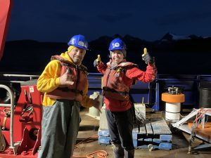 Two people stand next to each other wearing life vests. One of them is giving thumbs up while the other is giving shakas. They are standing on the ship's deck. There are mountains in the background. They are well lit, but it appears to be night.