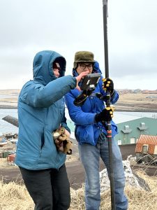 Harper Baldwin and Chris Tran stand next to each other. Chris is holding the Trimble instrument and they are both looking at the Trimble controller screen. They are outside on a hill. Behind them are various buildings and a large body of water. The sky is overcast.
