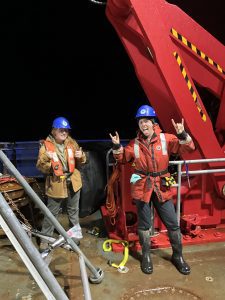 Two people stand next to each other wearing life vests. One of them is giving two thumbs up while the other is giving rock on hand signs. They are standing on a well lit deck at night. There is a large red metal crane next to them.