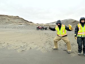 Chris Maio and Chris Tran stand on a sandy beach. Chris Maio is holding the field notebook with his arms out in a partial squat stance. There are two parked ATVs behind them and some small hills with dried grass are further back. The sky is overcast.