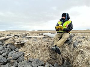 Chris Maio sits on driftwood looking at an open field notebook. The driftwood is on dried grass and surrounded by rounded basalt. The sky is overcast.