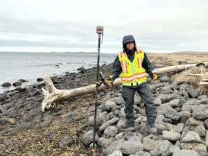 Chris Tran stands on vesicular rocks on a rocky beach. He is holding a Trimble instrument. The ocean water is to the left of the image and dried grass is to the right of the image.
