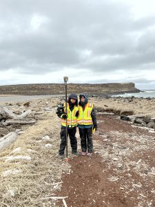 Chris Tran and Harper Baldwin stand next to each other on a mix of dried grass and dirt. Chris is holding a Trimble instrument. They are surrounded by driftwood and large rocks. The ocean can be seen behind them.