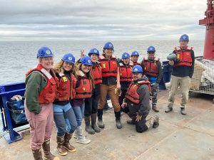 A group of people standing near each other for a group photo. They are standing on a deck. There is ocean behind them and the sky is overcast.