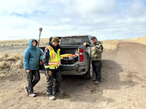 Harper Baldwin, Chris Tran, and Alex Zaochney standing around a parked truck. Harper is holding the Trimble instrument. They are standing on a dirt road with dried grass on either side. The sky is cloudy.