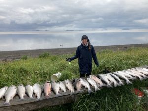 Harper stands behind a long row of fresh salmon. She is in tall grass. There is a beach in the background with calm, glassy waters. The sky is overcast.