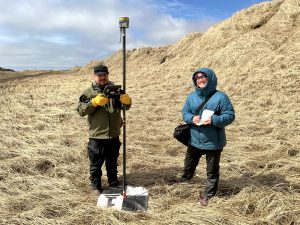 Alex Zaochney and Harper Baldwin stand next to each other. Alex is holding the Trimble instrument on a checkered tarp and Harper is holding the field notebook. They are standing in a field of dried grass. The sky is cloudy. 