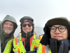 Chris Maio, Harper Baldwin, and Chris Tran stand next to each other for a selfie. There is water behind them. The sky is overcast. 