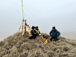 Three people sit on a hill with dried grass. Next to them is the Trimble base station. The sky is overcast. 