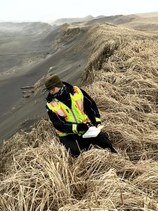 Chris Tran sits on the edge of a hill with dried grass. Down the slope of the hill is sand. Chris is holding a field notebook. The sky is overcast and misty.