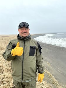 Alex Zaochney is standing with a thumbs up on a hill of dried grass that slopes down to a sandy beach. The sky is overcast. 