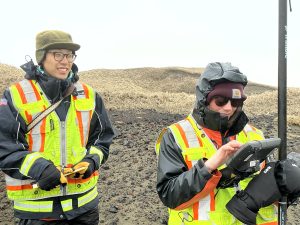 Chris Tran and Harper Baldwin stand next to each other on semi-rocky ground. Harper is holding the Trimble instrument and Chris is holding a field notebook. There are small hills with dried grass behind them and the sky is overcast. 