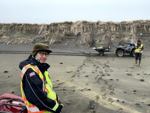 Chris Tran and Harper Baldwin are standing on a rocky beach near some ATVs. Harper is holding a Trimble instrument. There are bluffs further in the background with dried grass covering them. The sky is overcast. 
