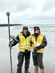 Chris Tran and Harper Baldwin stand next to each other on a sandy beach. Chris is holding a Trimble instrument and Harper is holding a field notebook. The sky is overcast.
