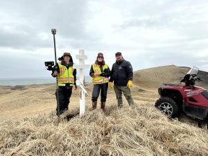 Chris Tran, Harper Baldwin, and Alex Zaochney stand on top of a hill with dried grass and a grave marker. An ATV is parked nearby. The sky is overcast and the ocean is visible in the distance. 
