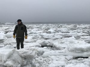 Alex Zaochney stands in a field of broken ice. Further back the ocean water is visible. The sky is overcast.