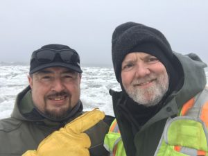 Alex Zaochney and Chris Maio stand next to each other for a selfie. Behind them is a field of broken ice. The ocean is visible further back. The sky is overcast. 